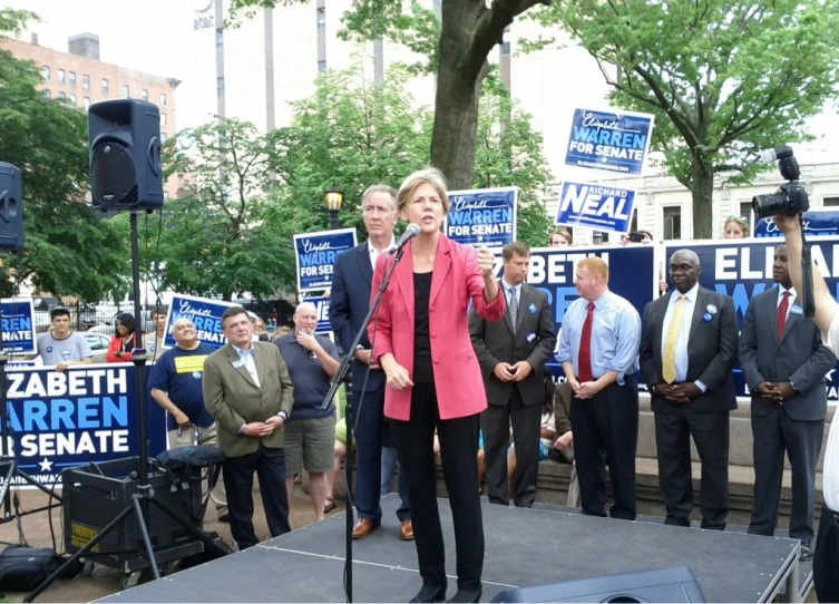 two women speaking into microphones at podium with people holding signs