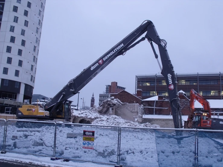 a crane stands in front of the edge of a construction site with a snowy building and other equipment in the background