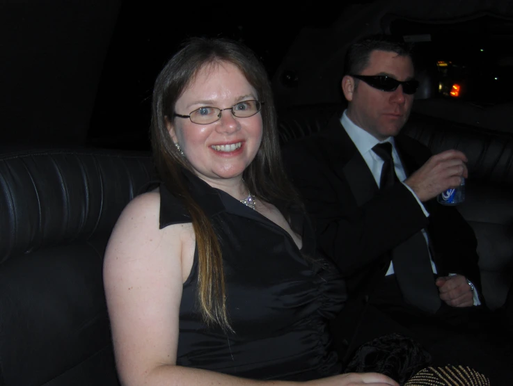 a woman with glasses smiling at the camera and a man in a suit sitting in the back of a vehicle