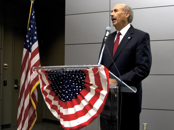 a man speaking at a podium with flags behind him