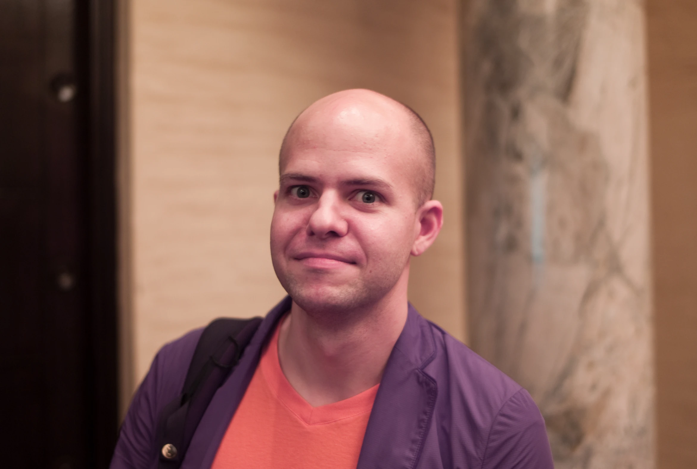 a balding man wearing an orange shirt standing in front of a pillar