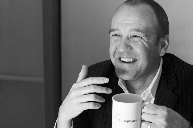 a man smiles and holds a cup while sitting at a table