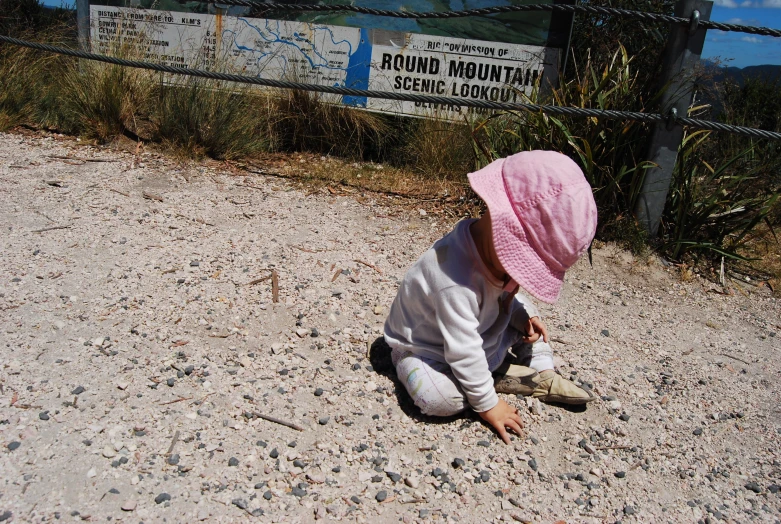 baby girl in pink hat playing on ground at fence