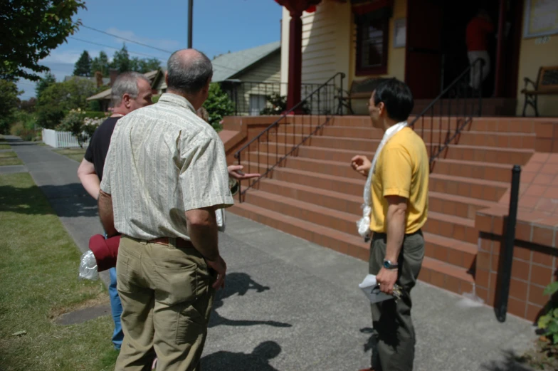 three men standing outside of a house one in yellow
