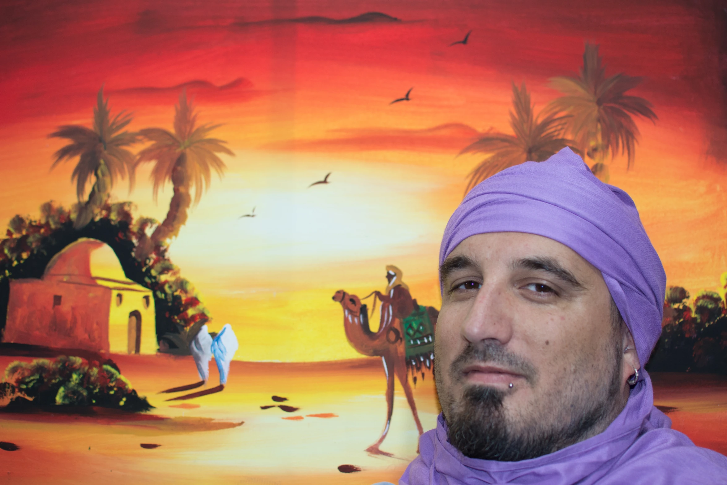 a man with a hat and purple scarf on standing in front of a painting of a sunset and three camels