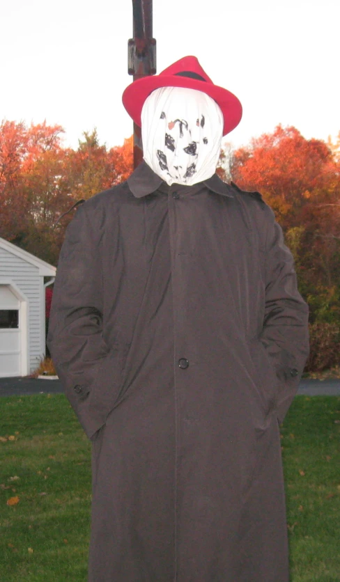 a man in a trench coat and hat standing in front of a cross with a face painted on it