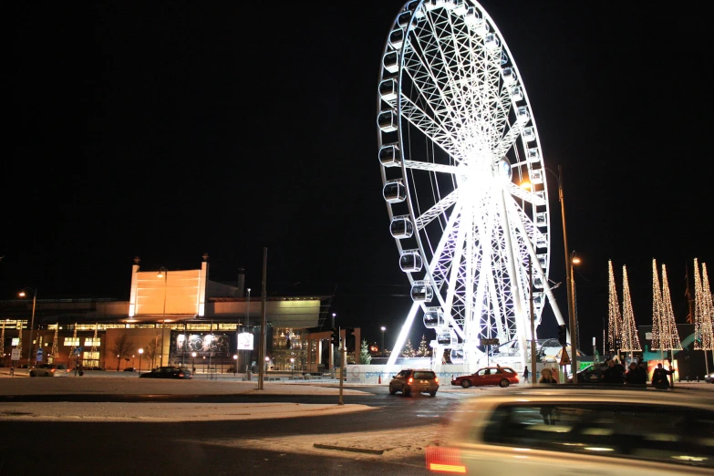 a carnival ferris wheel at night with people walking and a car passing by