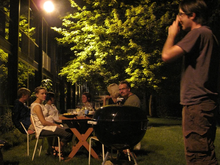 a man is talking on his cell phone while standing near people sitting at a table in the grass