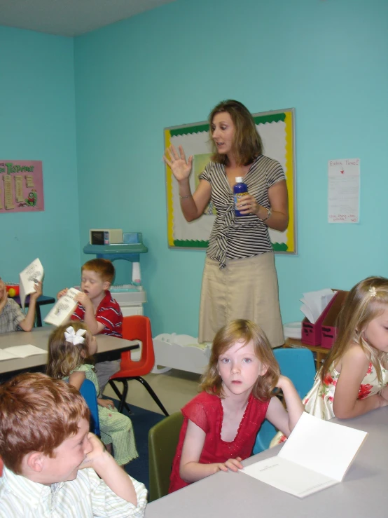 some young children sitting at desks and one woman stands