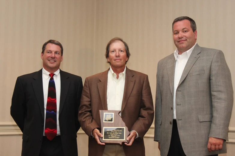 three men stand together while one man displays an award