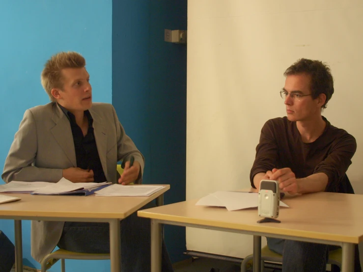 two people sitting at desks talking in front of a camera