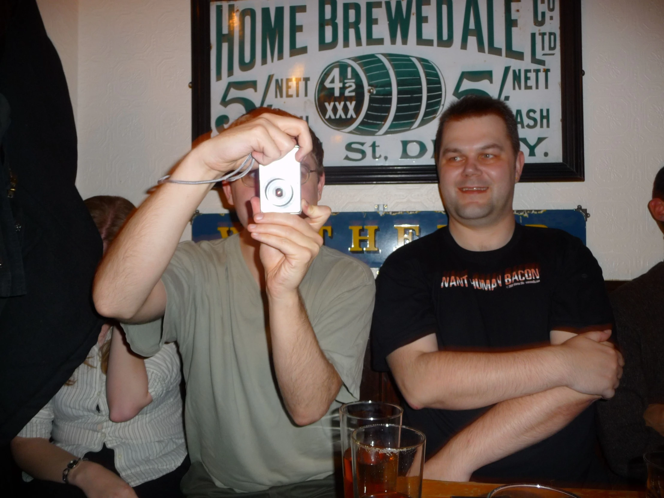 two men are sitting at a table with beer and one is holding up a camera