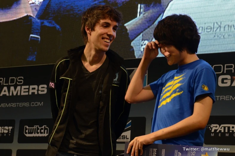 a young man smiles as he listens to another young person in a black shirt