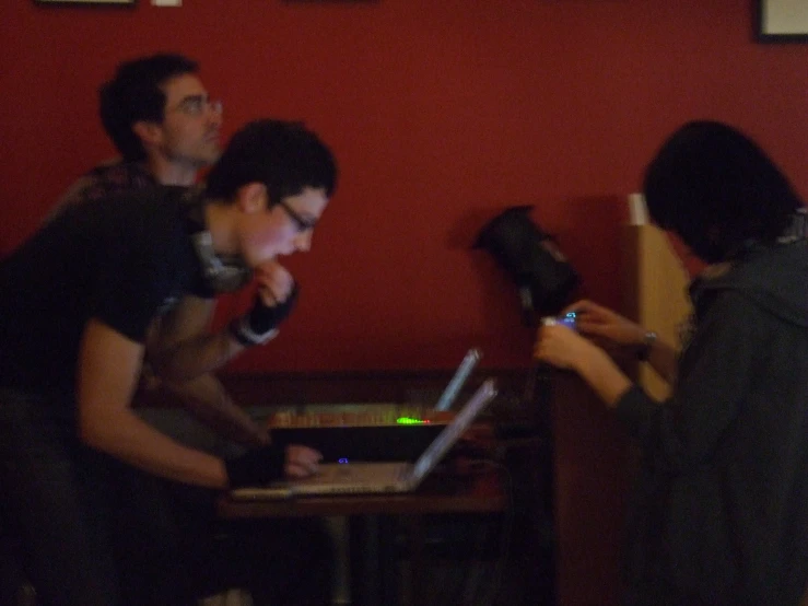 three men are gathered around a laptop in a dimly lit room