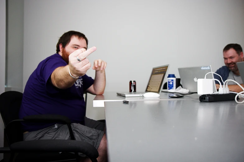 two men, one on his cell phone and the other in the office chair holding the same object up while sitting down at a table