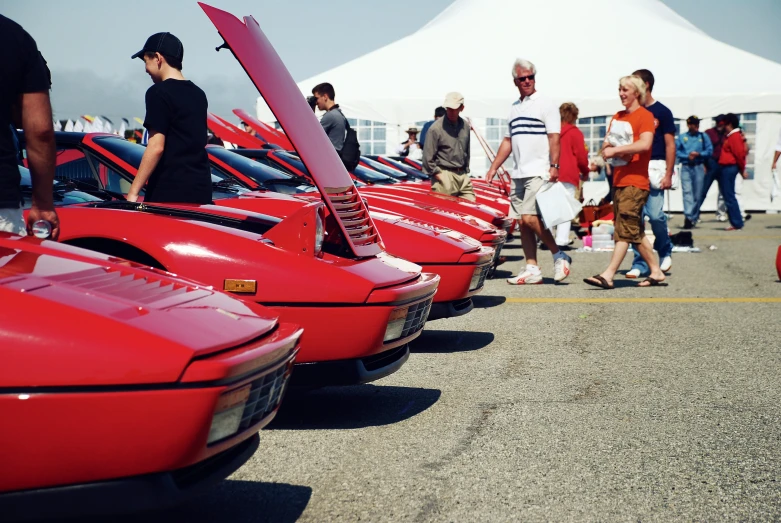 many people lined up near parked red cars in a lot