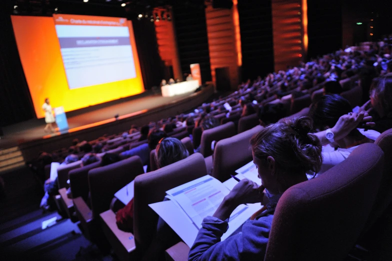 people in chairs at an auditorium watching a presenter on stage