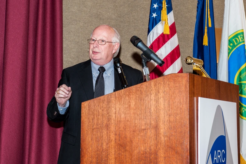 an old man speaking at a podium with flags and microphones