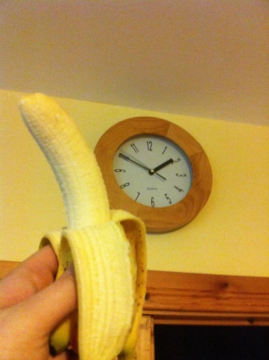 a hand is holding a ripe banana with the wall clock above