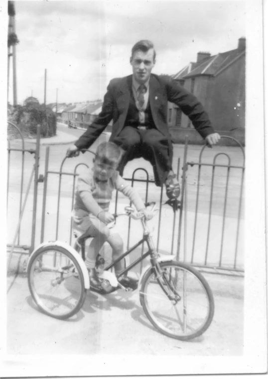 two boys in dress clothes on a bike, in front of a fence with an iron guard rail