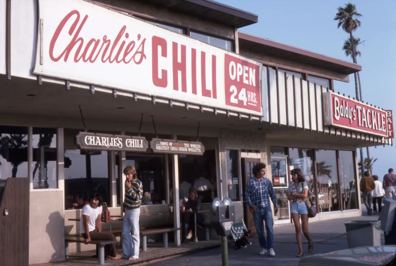 people walking by a deli with many signs on the front of it