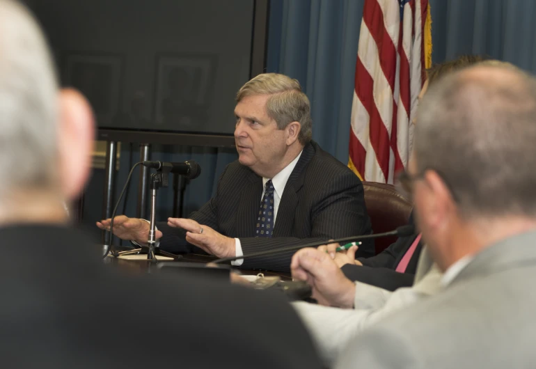 an older man in suit and tie speaks at a table with two other men and one is holding microphone