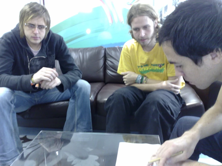 three men sit and talk around in front of a glass table