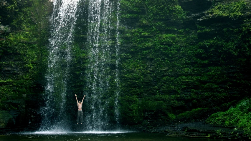 a woman is standing in front of a waterfall and raising her arms in the air