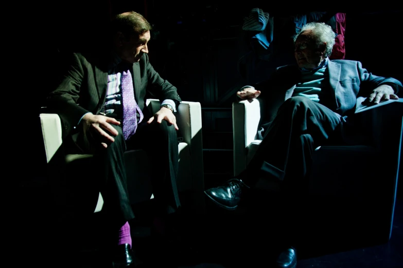 an older man sitting in white chairs wearing black suit