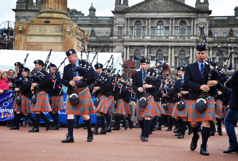 a parade of men in kilts and suits in uniforms with pipe bearer marching past an old building