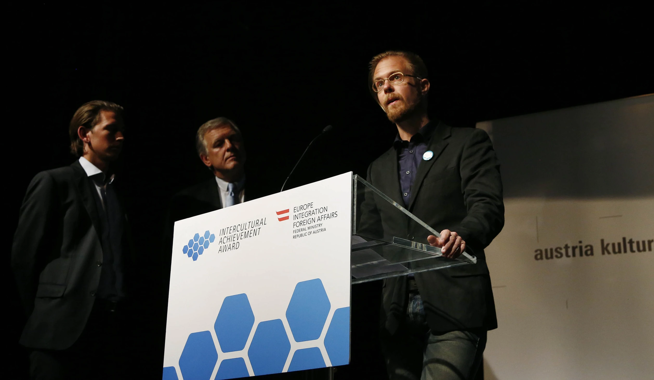 three men standing on stage at podiums while one man gives a speech