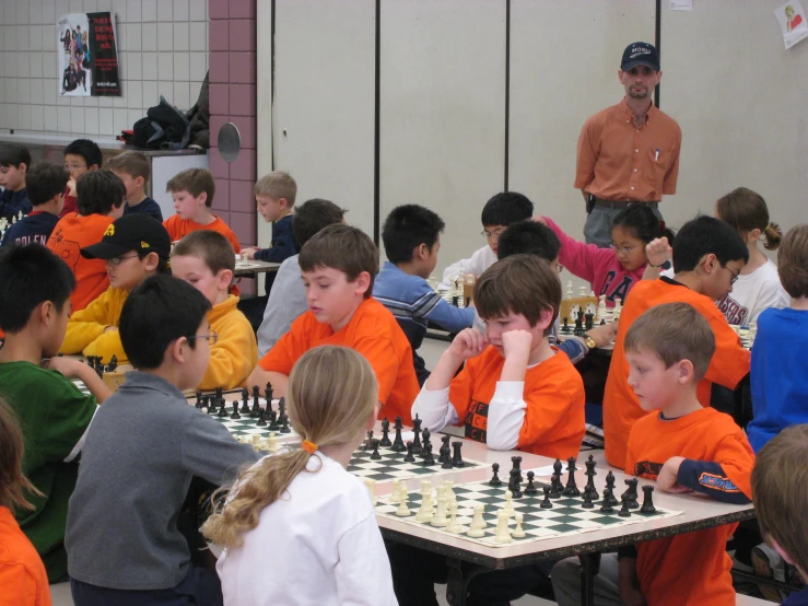 a man stands next to the back wall of a room of students in orange uniforms playing chess