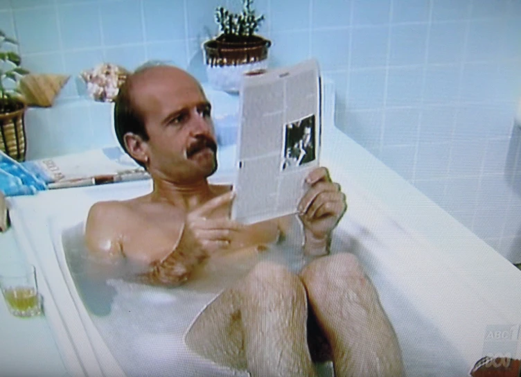 a man reading a newspaper on the edge of a bathtub