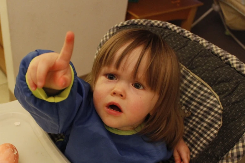 a  with her hand up is pointing at a piece of carrot in a high chair