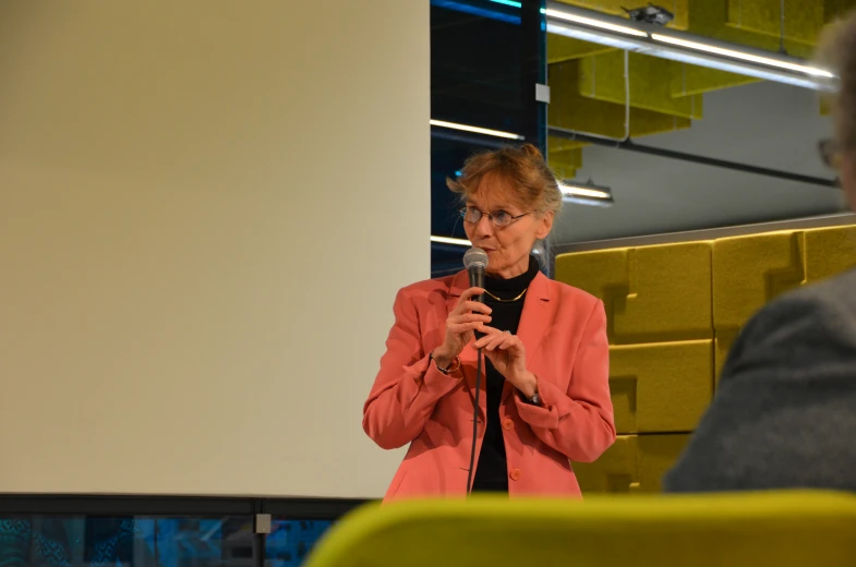 a woman with glasses and pink jacket speaking into microphone