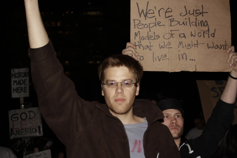 man holding up a placard that reads we're just people building made of a world to live in