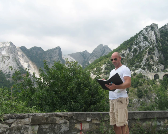 a man wearing a white shirt and tan shorts stands looking at a black folder while standing on the edge of a cliff