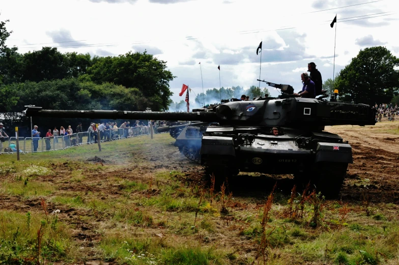 military tanks drive down a dirt road in front of spectators