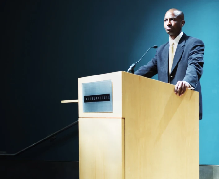 a man in a suit stands at a podium with his hands out