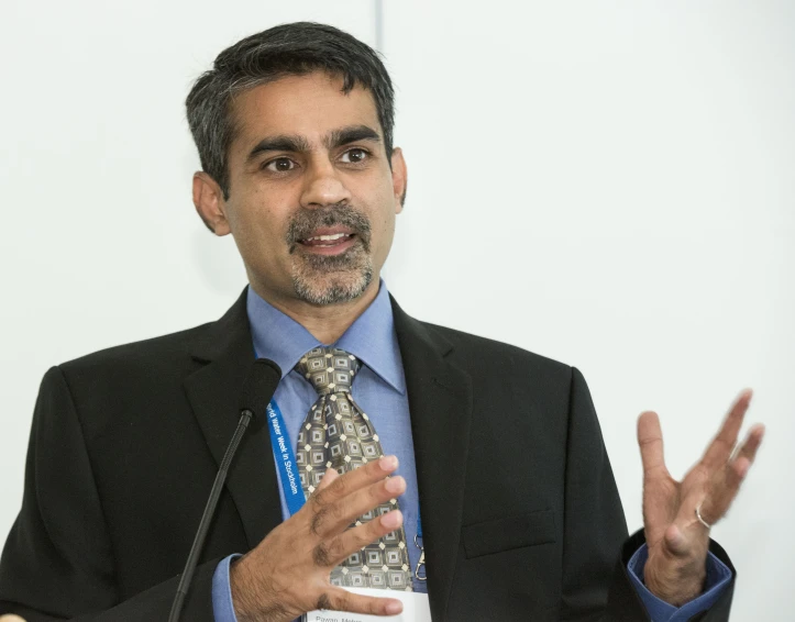 man wearing jacket and tie making gestures at podium