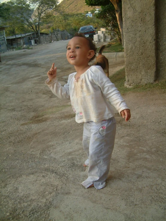 a baby standing on a dirt road wearing a hat and a white suit