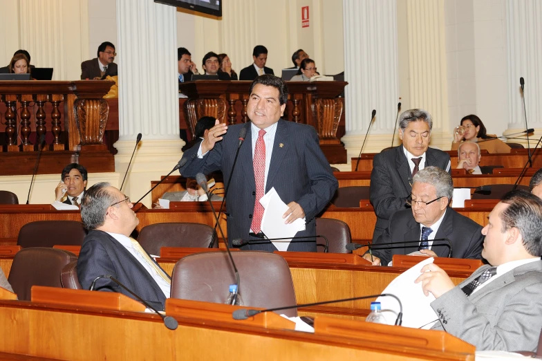 a man wearing a red tie standing behind microphones in a room with lots of people