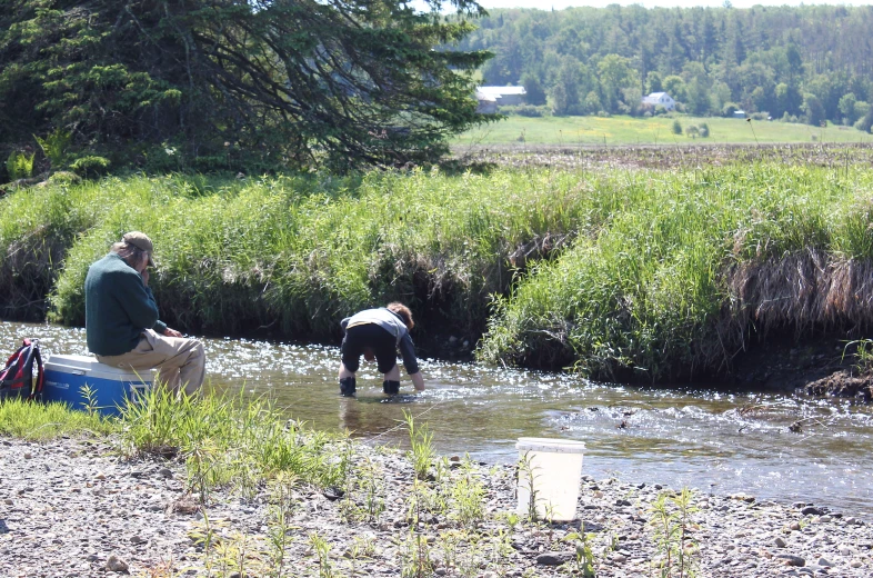 two people stand near a small river with their backs to the ground, while one holds a fish and another holds a book