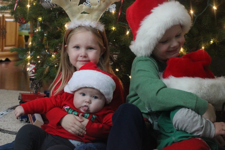 two girls, one holding a baby in a santa hat and one in a green top, sitting in front of a christmas tree