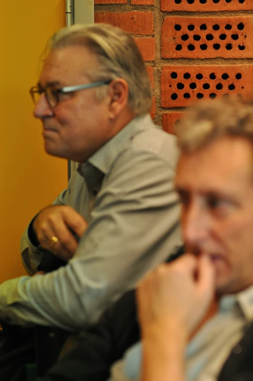 two men with glasses are seated looking at soing