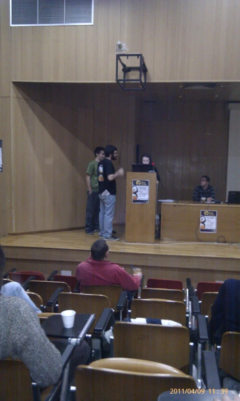 three people standing in a lecture hall, some siting and others stand with their back to the camera