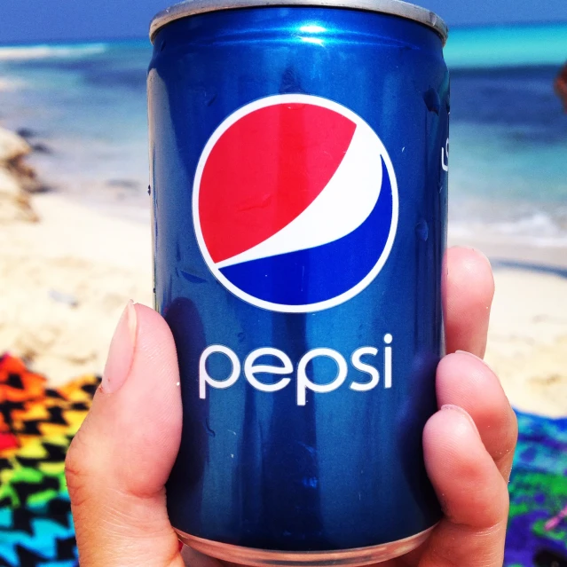 a pepsi can being held by a hand near the beach