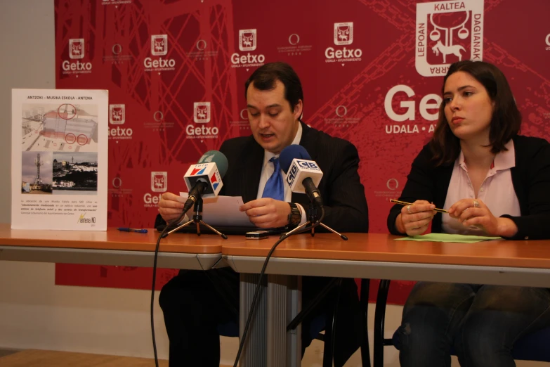 two people at a desk, one talking and the other holding microphones