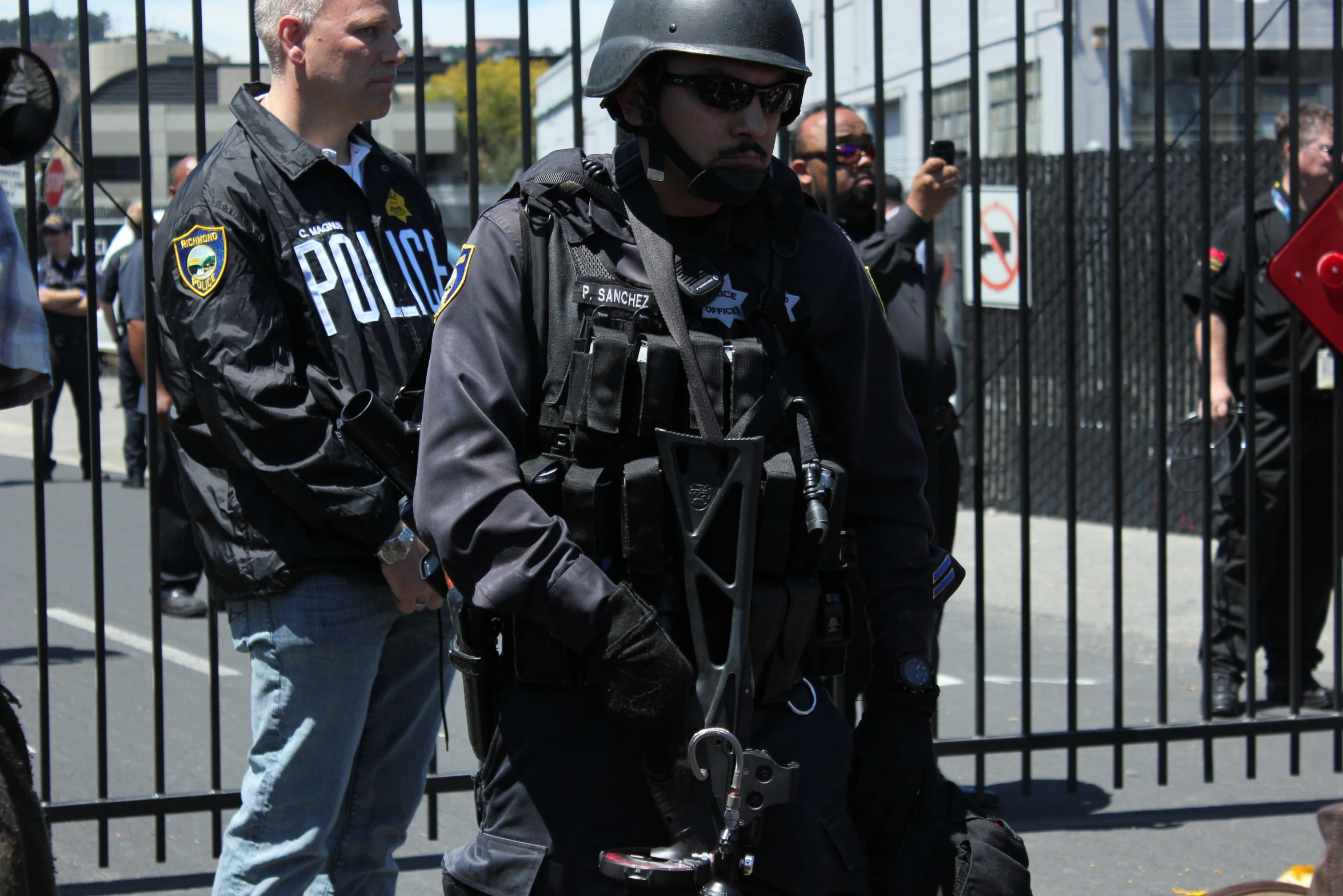 an armed police officer stands behind another man with bags