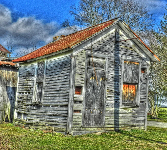 a rustic old gray house in a grassy field with trees and clouds in the background